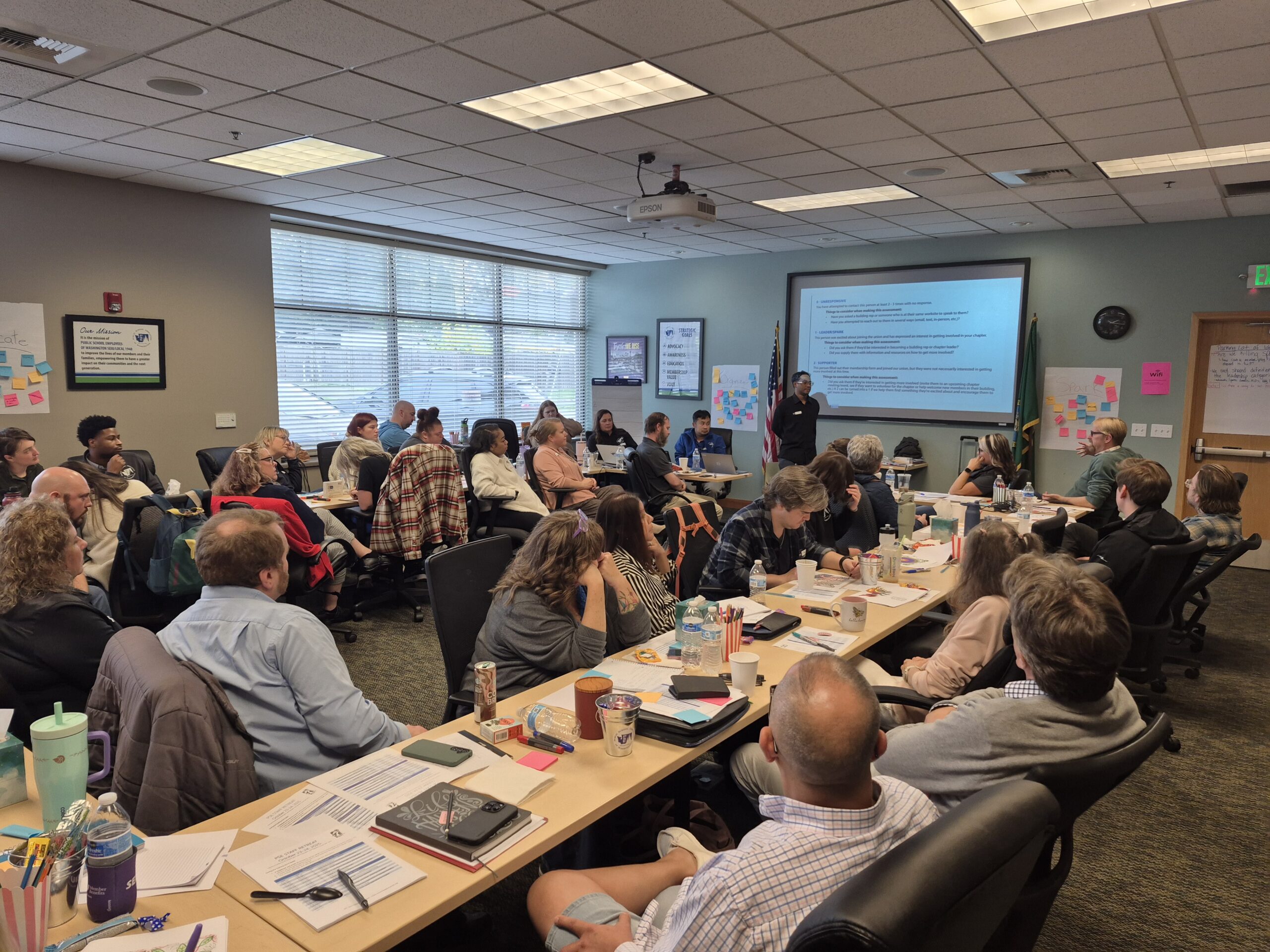 A large group of attendees seated around tables during a training session with an instructor presenting a slideshow at the front of the room